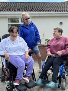 an image of two Paul's Place members sat in their wheelchairs in the garden. Our support worker is in between them and they are all laughing.