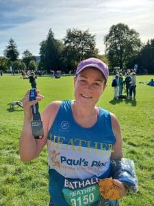 a picture of a lady who has just completed the Bath Half Marathon. She stands under sunny skies, is smiling holding up her medal and wearing a Paul's Place running vest. 