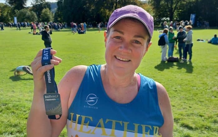 a picture of a lady who has just completed the Bath Half Marathon. She stands under sunny skies, is smiling holding up her medal and wearing a Paul's Place running vest.